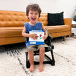 Toddler sitting in a FeedMe booster seat holding a book, in a living room setting.