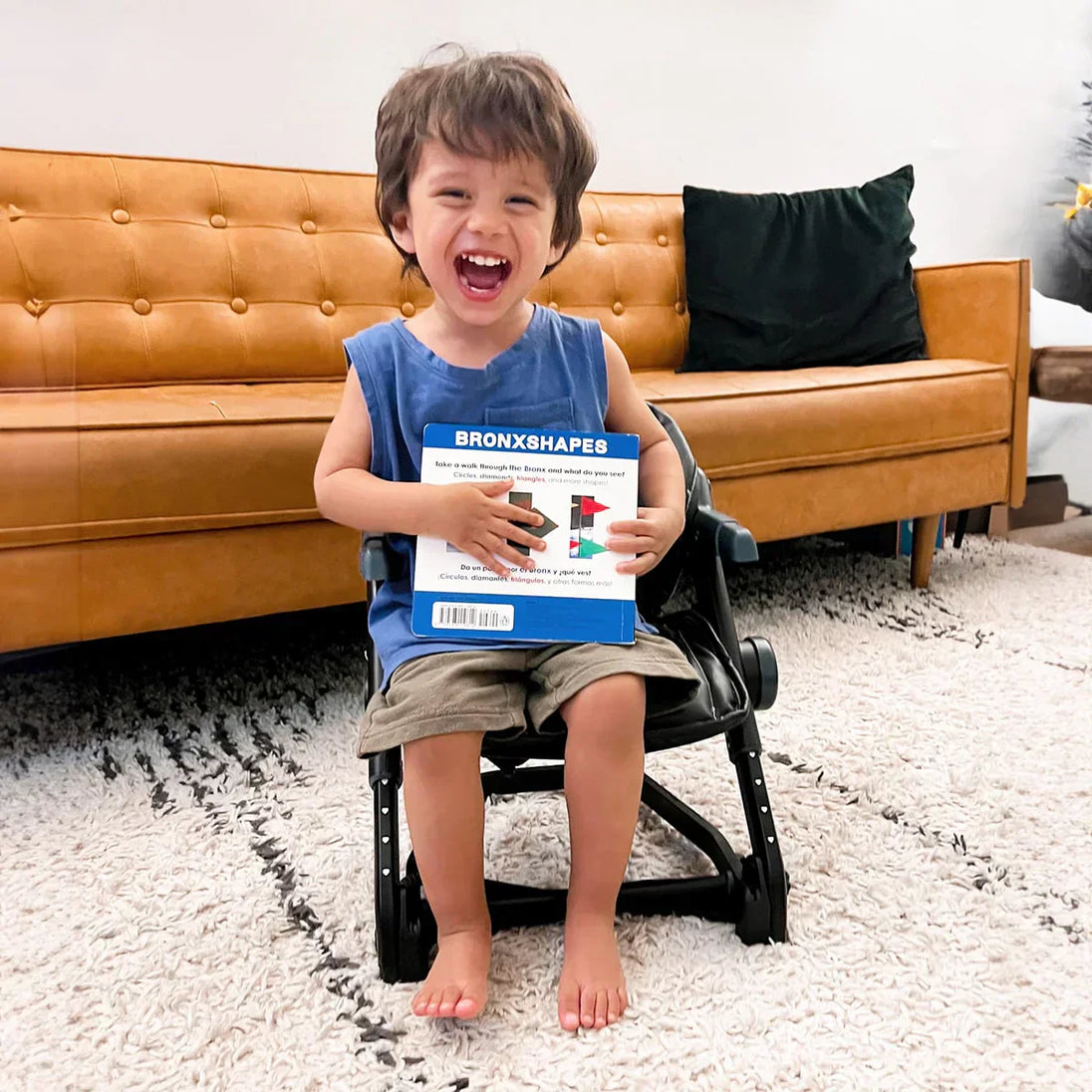 Toddler sitting in a FeedMe booster seat holding a book, in a living room setting.