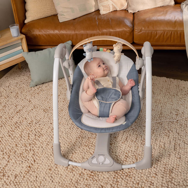 Baby enjoying Ingenuity Swing 'N' Go Portable Swing in Chambray with toy bar in cozy home setting.